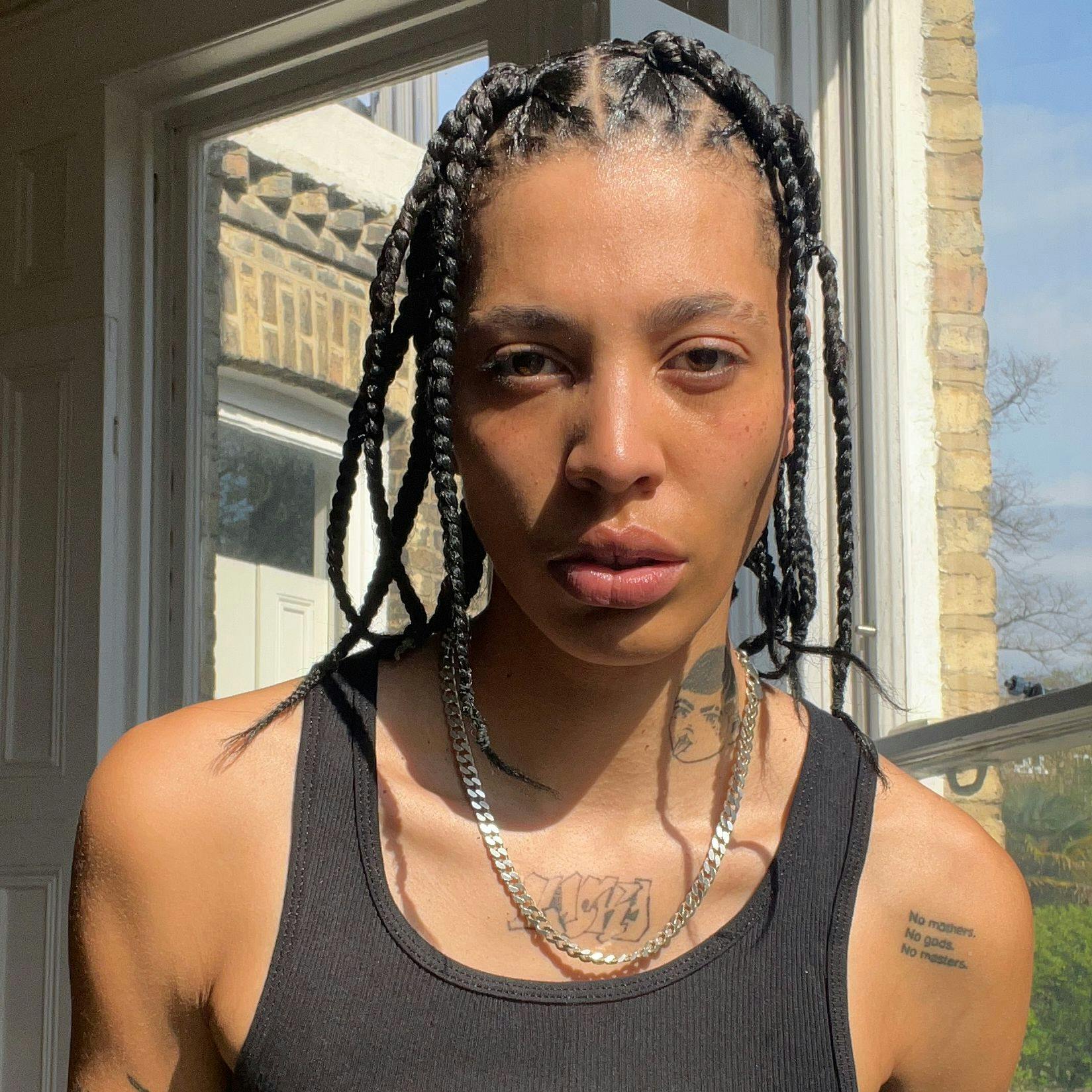 Kai facing the camera with soft shadows across their face, wearing a black tank top and silver chain, tattoos visible on their neck and arms.