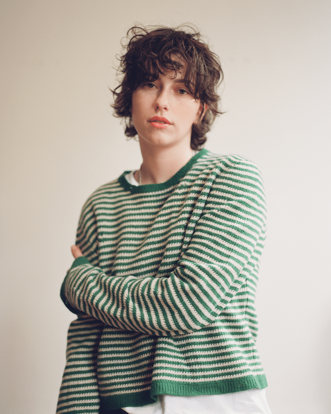 King Princess, aka Mikaela Straus, is staring at the camera with a soft but serious expression. They're wearing a green and white striped sweater, standing with one arm wrapped around their stomach. . 