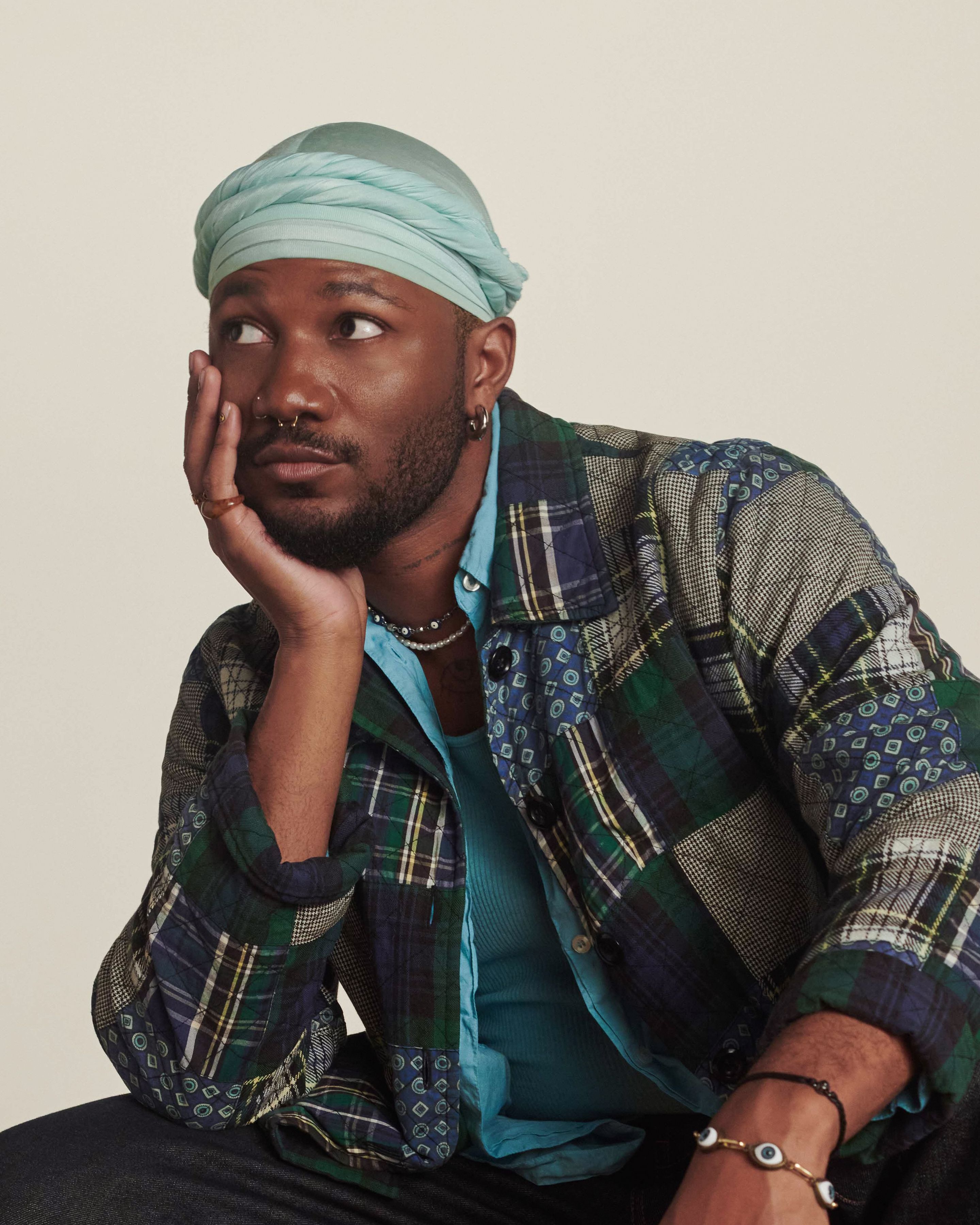 Shahem Mclaurin, relaxed and seated with a thoughtful expression looking to the side. They're wearing a patch-work jacket, seated in front of a tan background.