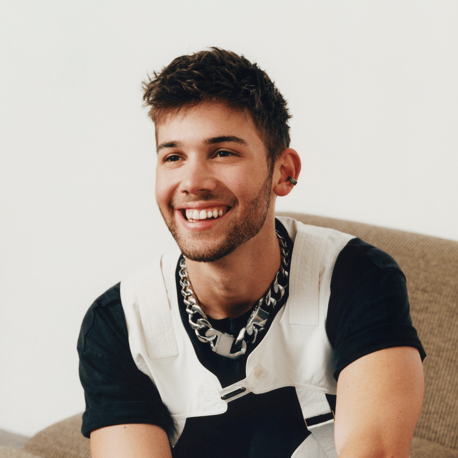 A portrait of Grant Knoche, against a white brick wall, is looking up into camera with a gentle smile in a light blue fleeced polo.