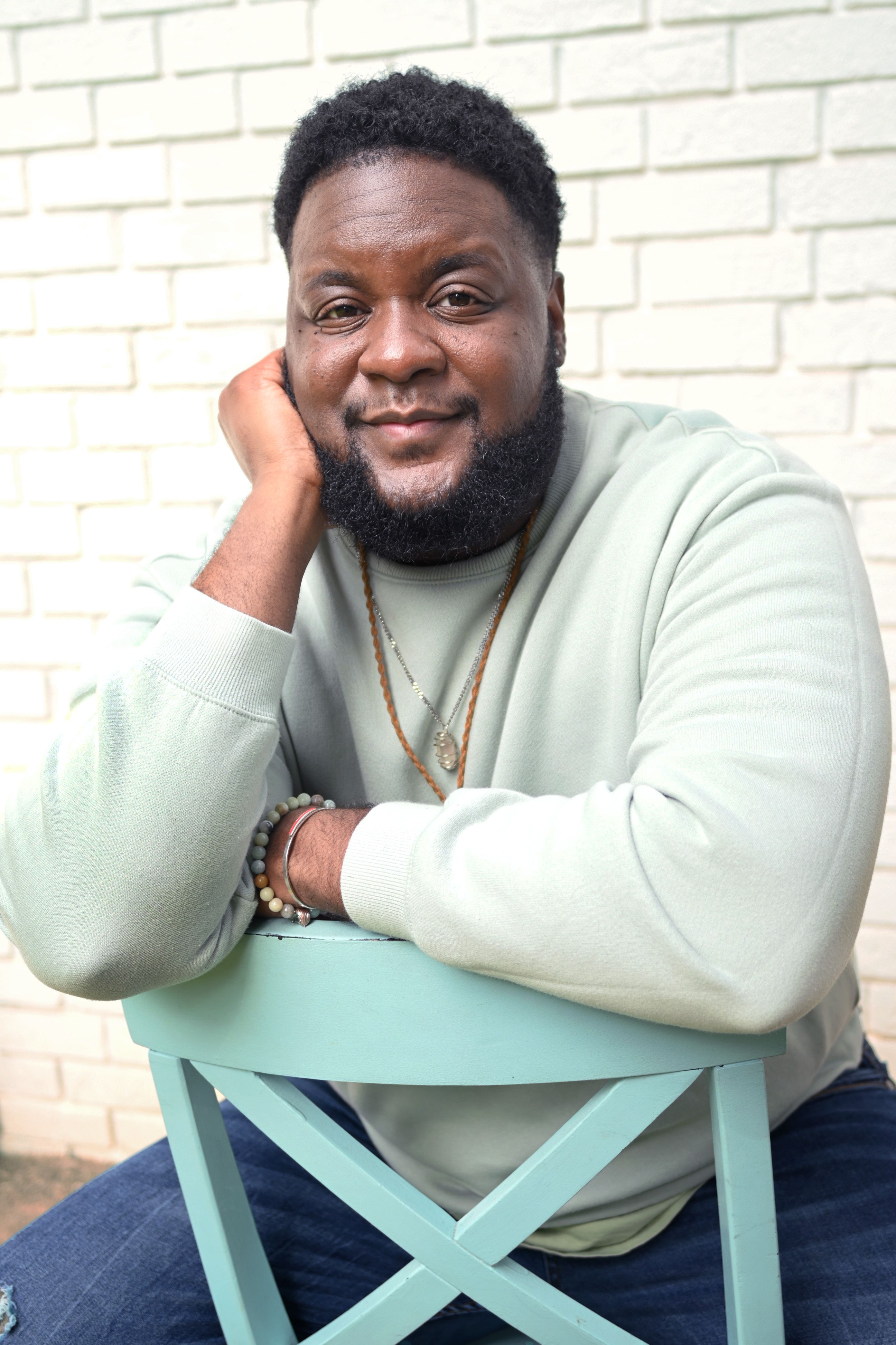 Moe Ari Brown sits on a backwards chair in front of a white brick wall