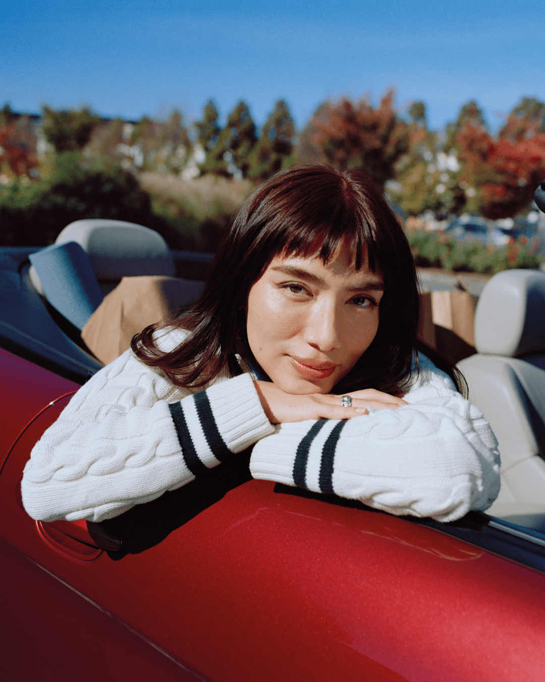 Ash is leaning on a car door, looking at the camera on a sunny day.