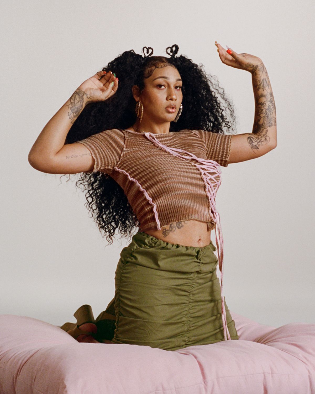 Bay posed, lips slightly pursed, kneeling on a pink floor cushion against a green and white backdrop. She wears a short sleeved brown and tan striped shirt, her tattoos are visible on her arms.