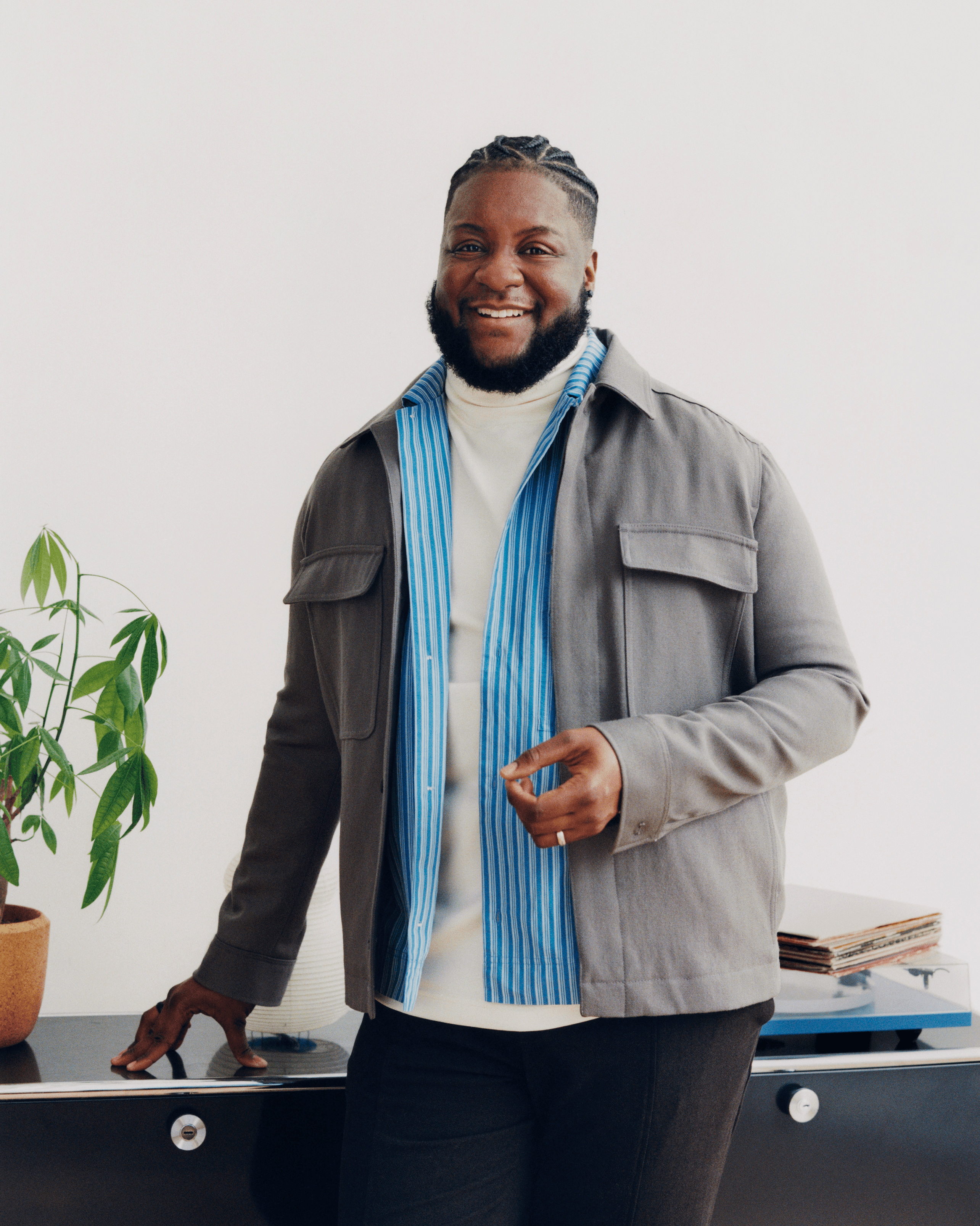 A shoulder-up portrait of Moe Ari Brown, wearing a light denim washed button up. They have a dazzling smile up against a marble background.