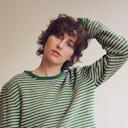 King Princess, aka Mikaela Straus, is staring at the camera with a soft but serious expression. They're wearing a green and white striped sweater, standing with one arm bent up toward their head. 