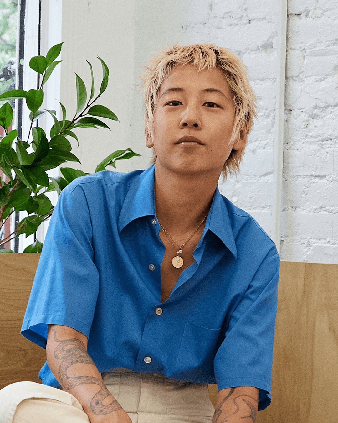 A portrait of Masami sitting, looking straight into to camera. With a soft smile and their head slightly upturned, they express an inviting confidence.