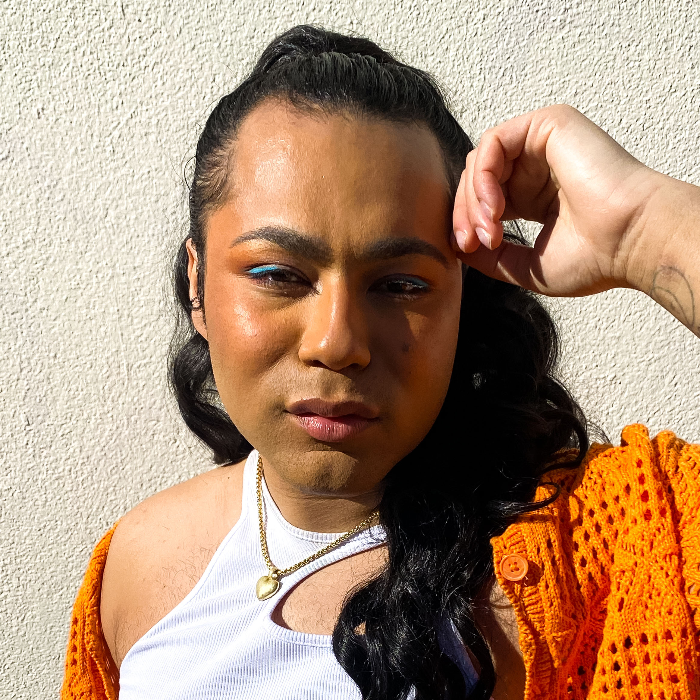A portrait of Travis looking into the camera, head resting on one arm, wearing a white tank and an orange cardigan