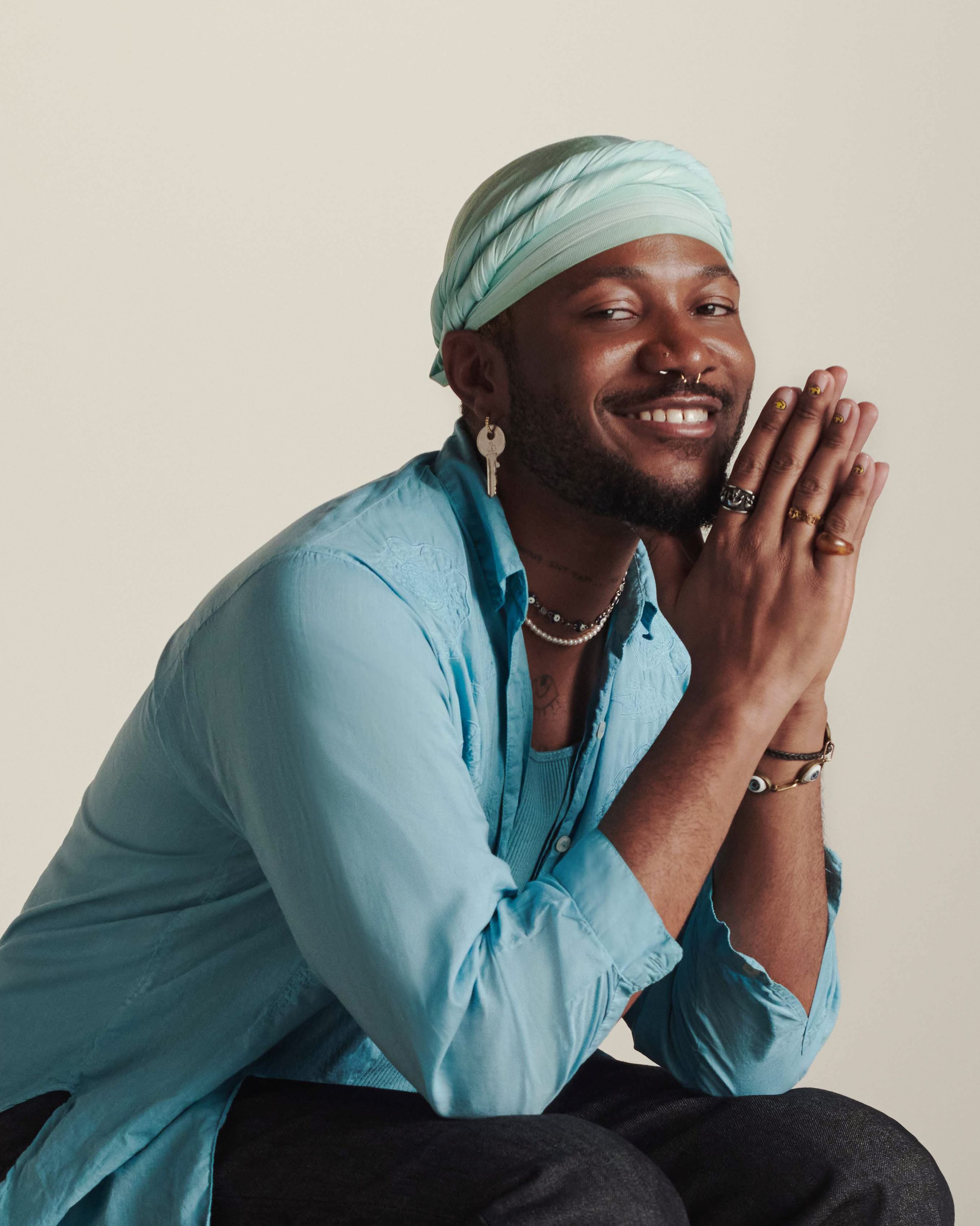 Shahem Mclaurin, hand resting on cheek, looking into the camera, clouds reflecting in the windows of the building behind them.. They're wearing a black sweater with yellow smiley faces stitched in.